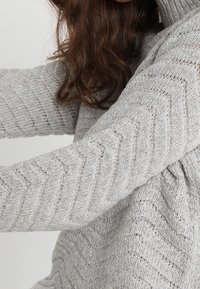 Hellgrauer gestrickter Pullover mit Chevron-Muster, hohem Kragen und langen Ärmeln. Die Textur wirkt weich und gemütlich und zeigt einen geschichteten Look.