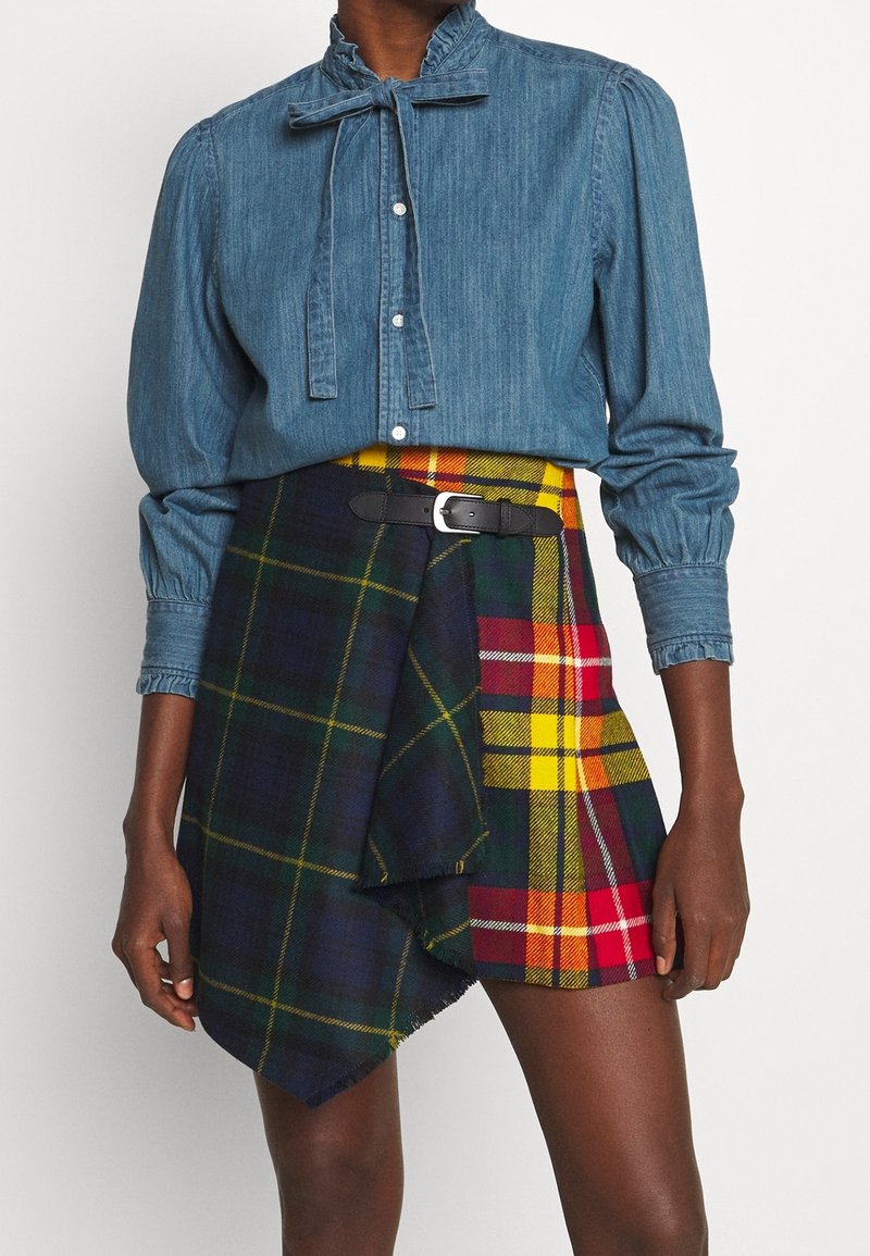 Personne à la peau foncée portant un chemisier en denim bleu à manches longues avec col à volants et une jupe asymétrique en tartan multicolore avec une ceinture noire.
