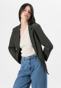 Una modelo posa con confianza, vistiendo un blazero verde oscuro sobre un suéter claro y Jeans de tiro alto azules, contra un fondo blanco liso.