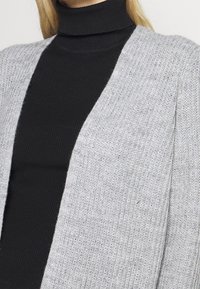 Szary sweter na drutach z otwartym frontem, o prążkowanej fakturze, noszony na dopasowanym czarnym golfie.