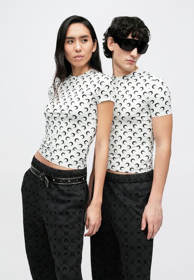 Camisetas blancas con un estampado de media luna negra, combinadas con pantalones negros que tienen un diseño sutil. Los accesorios del modelo incluyen gafas de sol y un cinturón.