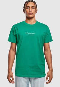 Camiseta de algodón verde con texto en árabe e inglés "Ámate a ti mismo primero". Con cuello redondo clásico y mangas cortas, tiene un corte relajado.