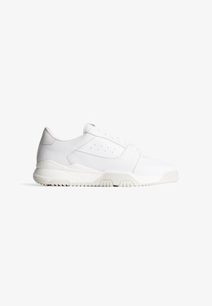 Weißer Sportschuh mit einem strukturierten Lederobermaterial, abgerundeter Zehenform und einer dicken Gummisohle mit einem geriffelten Muster für besseren Halt.