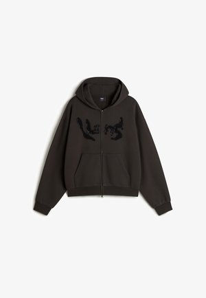 Mørkegrå zip-up hoodie med en rummelig hætte, to frontlommer og et tekstureret, skinnende sort logo på tværs af brystet. Blød stoffinish.