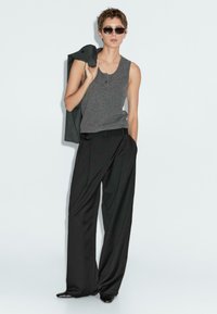 Top grigio senza maniche, pantaloni neri a vita alta e scarpe nere aderenti. La modella tiene un giubbotto scuro ed indossa occhiali da sole neri. Texture lisce.