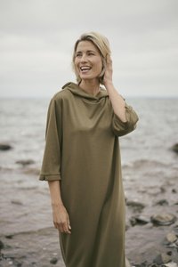 Robe à capuche vert olive en tissu doux, dotée de manches courtes et d'une coupe décontractée. Portée par une femme près d'un rivage rocheux.