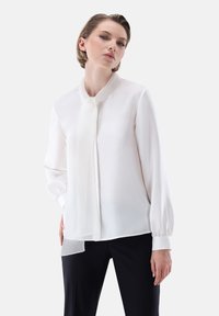 Blusa bianca in chiffon con colletto standing, maniche lunghe e dettaglio al collo annodato. Presenta una texture liscia e leggere trasparenze all'orlo.