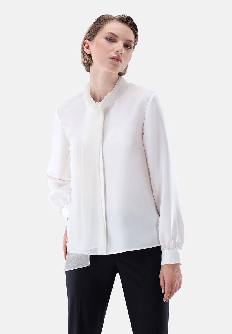 Blusa bianca in chiffon con colletto standing, maniche lunghe e dettaglio al collo annodato. Presenta una texture liscia e leggere trasparenze all'orlo.