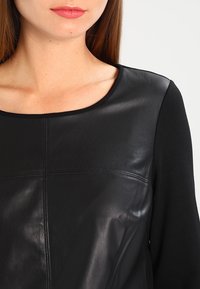 Schwarzes Oberteil mit einer glatten Lederfront und weichen Stoffärmeln, einem abgerundeten Halsausschnitt und dezenten Nahtdetails.