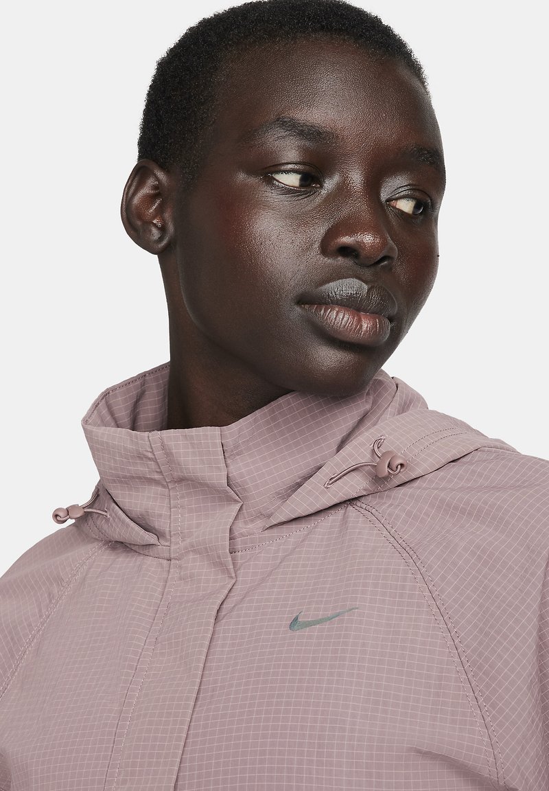 Close-up van een persoon in een mauve Nike-jack met een capuchon en trekkoorden, die naar de zijkant kijkt tegen een effen achtergrond.