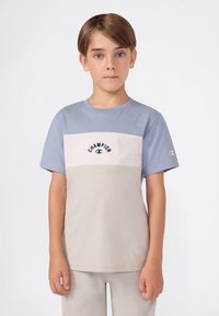 Niño con cabello corto castaño, vistiendo una camiseta color bloque de la marca Champion en azul y beige, y pantalones beiges, de pie contra un fondo blanco.