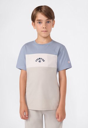 Niño con cabello corto castaño, vistiendo una camiseta color bloque de la marca Champion en azul y beige, y pantalones beiges, de pie contra un fondo blanco.