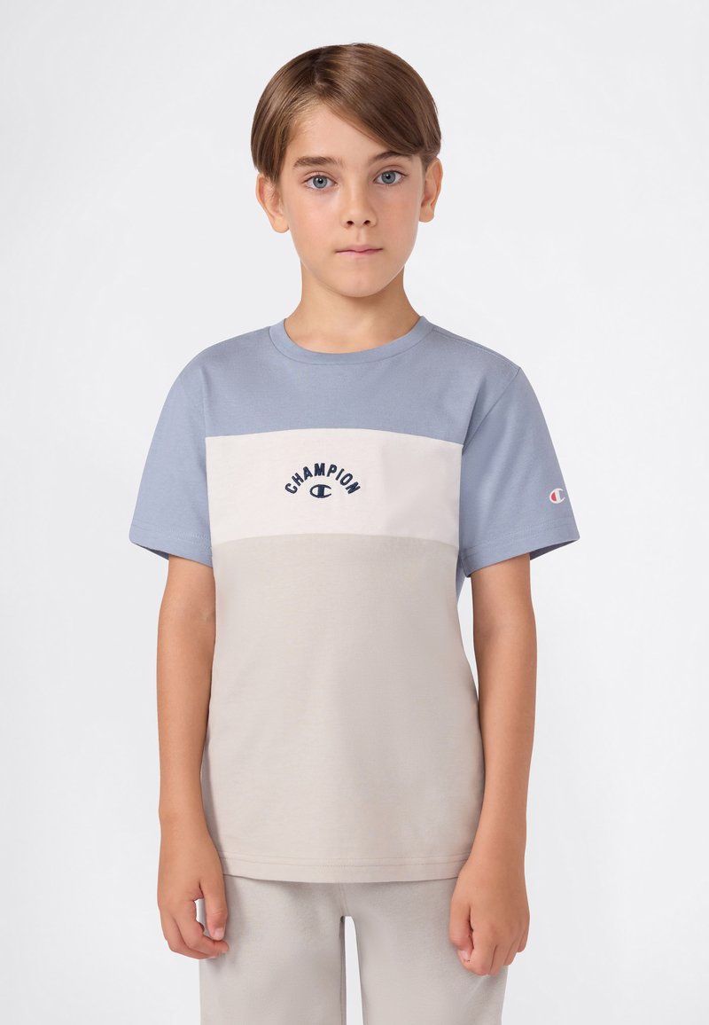 Niño con cabello corto castaño, vistiendo una camiseta color bloque de la marca Champion en azul y beige, y pantalones beiges, de pie contra un fondo blanco.