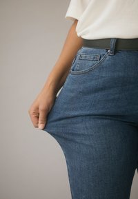 Dżinsy z denim w średnim niebieskim odcieniu z klasycznym pięciokieszeniowym krojem. Ręka ściskająca materiał, pokazująca lekki rozciągnięcie, widoczne szwy.