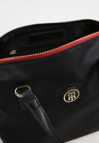 Sort håndtaske med guldfarvet dobbelt lynlås, rød indvendig lynlås, læderrem og cirkulært metallisk logo-emblem på forsiden.