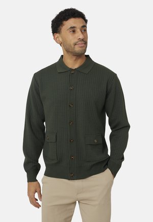 Man draagt een donkergroen gebreid vest met knopen, kraag en twee voorzakken, gecombineerd met beige broek, kijkt iets naar rechts.
