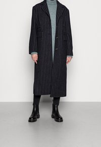 Námořnický pruhovaný kabát s klopovým límcem, dvěma předními kapsami a zapínáním na knoflíky, doplněný světle modrým svetrem a černými kotníkovými botami.