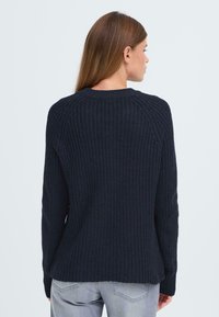 Pull en maille bleu marine avec une finition texturée, col rond et poignets ribbed. Présente une coupe légèrement ample et un dos ouvert.