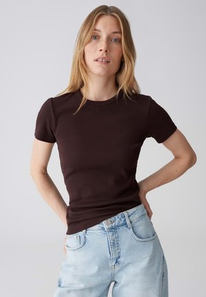 Frau mit blondem Haar, die ein dunkelbraunes kurzärmeliges Oberteil und hellblaue Jeans trägt, steht mit einer Hand in der Hüfte vor einem schlichten Hintergrund.