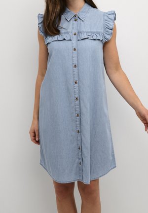 Světle modré džínové šaty bez rukávů s knoflíky vpředu, volánkovými rameny a límcem, nošené osobou na jednobarevném pozadí.