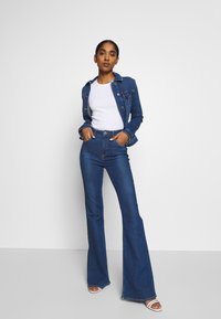 Jeansjacka och högmidjade utsvängda jeans i mörkblått. Vit åtsittande t-shirt under. Skorna är öppna vid tårna med en rem. Slät textur.