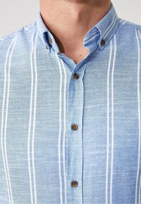 Camicia a righe blu chiaro con colletto button-down, caratterizzata da strisce verticali bianche a contrasto e bottoni marroni. Tessuto texturizzato.