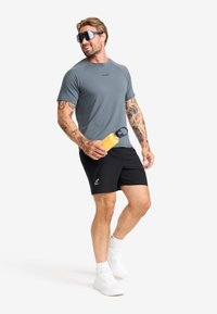 Athletic outfit featuring a gray short-sleeve shirt, black shorts, and white sneakers. Holding a yellow water bottle, with sunglasses accentuating the look.