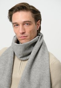 Grå uldhalstørklæde svunget om halsen, med en blød tekstur og en forlænget, rektangulær form. Enkel design uden mønstre.