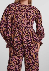 Haut à imprimé léopard avec manches ballon, détails smockés et col montant. Le tissu présente des tons marron, noir et fuchsia. Assorti d'un pantalon coordonné.