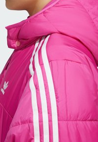 Růžová péřová bunda s vysokým límcem, se zabudovanými bílými pruhy po stranách a malým logem na hrudi. Textura se zdá být hladká a polstrovaná.