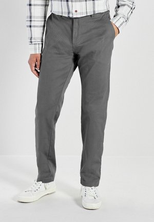 Homme portant un pantalon gris, des baskets blanches et une chemise à carreaux blanche, debout avec une main dans la poche contre un fond uni.