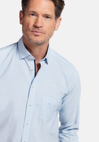 Lichtblauw overhemd met kraag, enkele borstzak en subtiele textuur. Heeft witte knopen en schone lijnen.