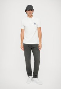 Pólo branco com um gráfico de animal azul, combinado com calças cinza-escuras e tênis brancos. Os acessórios incluem um boné escuro.