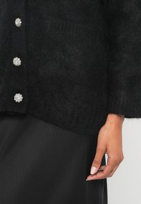 Μαύρη χνουδωτή ζακέτα με πέντε κουμπιά στολισμένα με λουλούδια, με άνετη εφαρμογή, βαθιές τσέπες και ριμπ μανσέτες. Φαίνεται απαλής υφής.