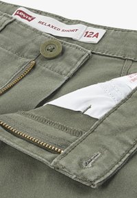 Shorts décontractés vert olive en coton, dotés d'une fermeture éclair en laiton, d'un bouton-pression et d'une étiquette intérieure en tissu blanc.