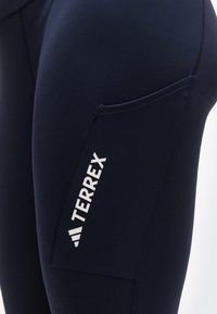 Κοντινή λήψη από σκούρο μπλε αθλητικό κολάν με πλαϊνή τσέπη που φέρει το κάθετο λογότυπο «TERREX» σε λευκό χρώμα.