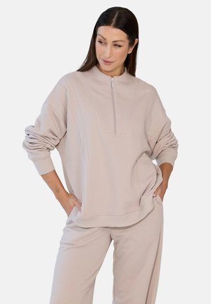 Kobieta z długimi ciemnymi włosami stoi z rękami w kieszeniach, ubrana w jasnobeżową bluzę rozpinaną na zamek oraz pasujące spodnie na białym tle.