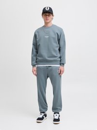 Pelēks sporta krekls un atbilstošas bikses, izgatavoti no mīkstas auduma, ar mazu logo uz krūtīm un biksēm. Melna cepure un sporta apavi noslēdz šo izskatu.