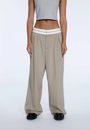Person trägt beige weite Hose mit weißem Bund und schwarzen Schuhen, kombiniert mit einem grauen, kurzen ärmellosen Oberteil.