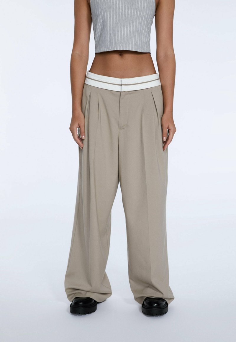 Personne portant un pantalon beige à jambes larges avec une ceinture blanche et des chaussures noires, associé à un top court gris sans manches.