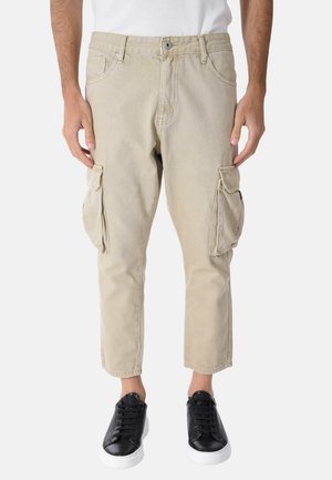 Uomo con pantaloni cargo beige corti con tasche con patta e sneakers nere basse con suole bianche.