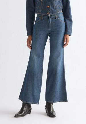 Femeie purtând blugi albaștri cu talie înaltă și pantaloni evazați, și cizme negre cu vârf ascuțit, mâinile relaxate pe lângă corp, pe un fundal simplu.