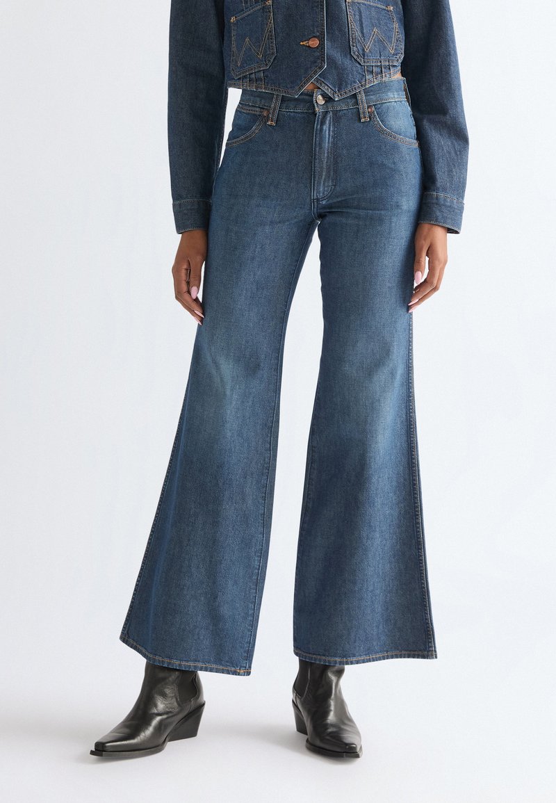 Femme portant un jean large taille haute bleu et des bottines noires à bout pointu, les mains détendues le long du corps, sur un fond uni.