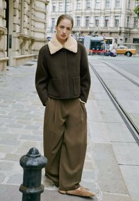 Veste en laine marron avec un col en fausse fourrure beige, associée à un pantalon large marron. Scène située dans une rue de la ville avec des pavés.
