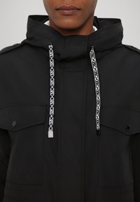 Veste noire avec capuche, dotée de cordons de serrage blancs à motif, de deux poches avant et de fermetures à boutons-pression. Texture lisse et tissu durable.