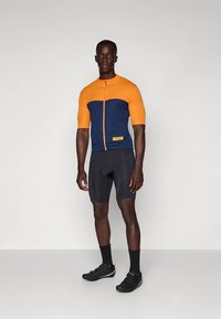 Kortärmad cykeltröja i orange och marinblå, med en dragkedja framtill, kombinerad med svarta shorts och cykelskor.