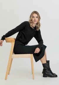 Musta neulepaita ja vihreät hame istuvalle hahmolle, jossa on puinen tuoli, jossa on kudottuja yksityiskohtia. Mustat nilkkurit, joissa on metallikoristeita.