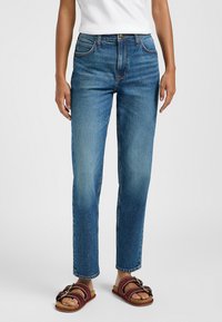 Jeans de mezclilla azul de pierna recta con una cintura de media altura, diseño de cinco bolsillos y un desvanecimiento sutil. Combinados con sandalias burdeos de tiras.