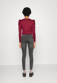 Mujer con el cabello corto y rizado, llevando una blusa burgundy de mangas abullonadas, jeans ajustados grises, calcetines transparentes con lunares y mocasines negros, de espaldas.