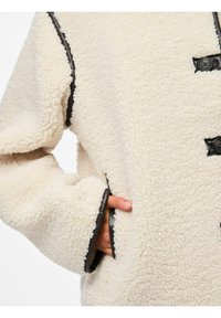 Kremfarget fleecejakke med teksturert overflate, med svarte kontrastkanter og knapper, samt sidelommer med en synlig hånd.